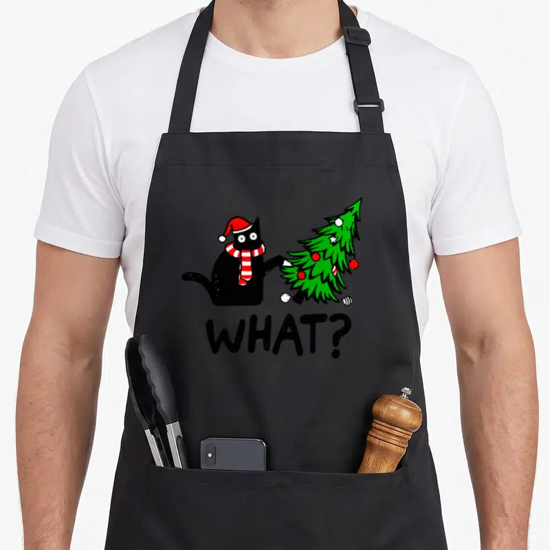 Pushing black cat apron - Cartoon black cat in Santa hat pushing green Christmas tree with text WHAT? — shown on a black cotton canvas apron against a white background