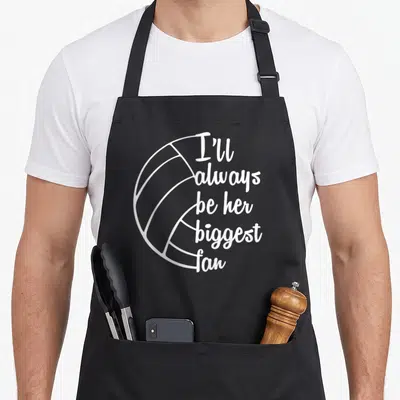 Coach volleyball dad apron - White line art volleyball with stacked text I'll always be her biggest fan — shown on a black cotton canvas apron against a white background