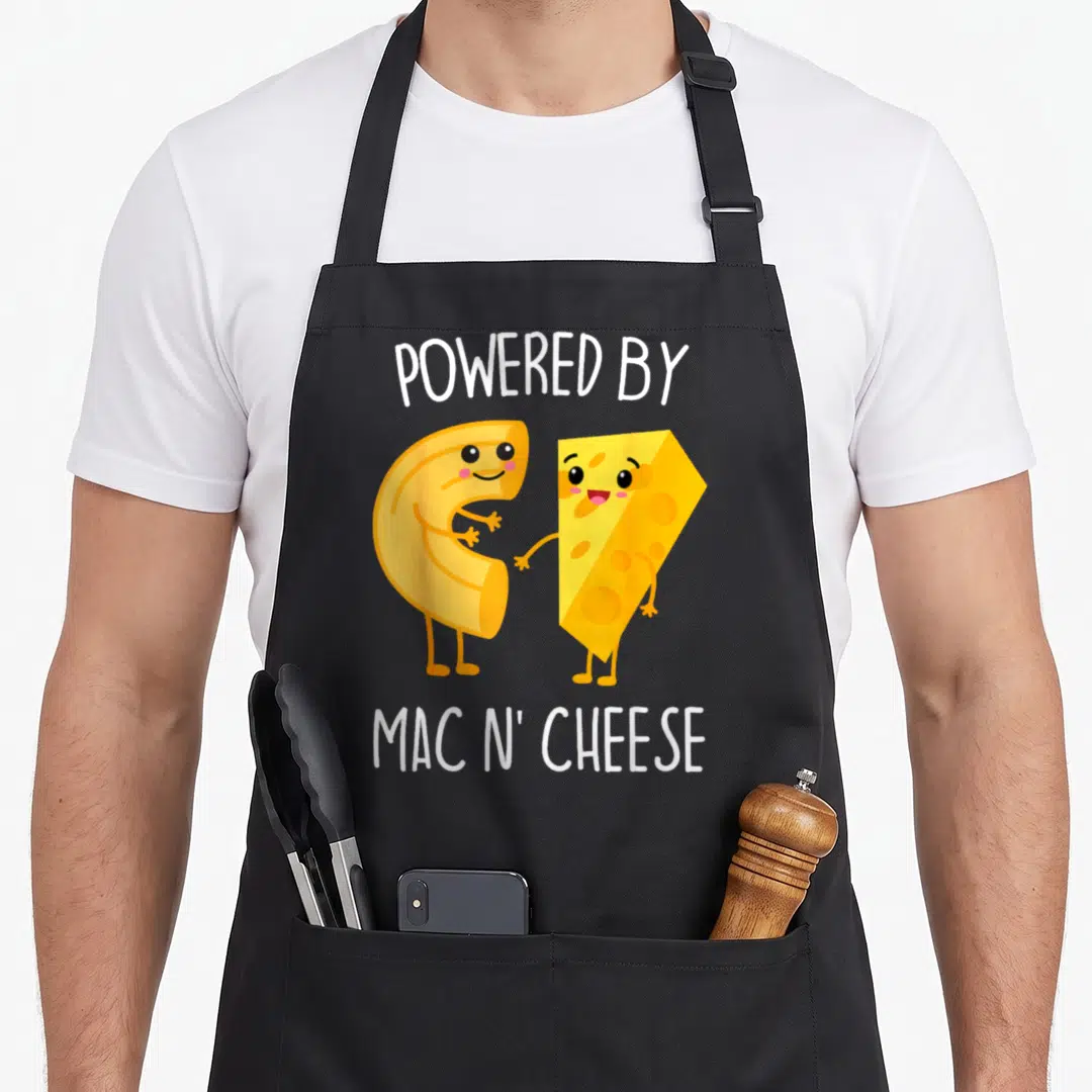 Powered mac apron - Cartoon macaroni and cheese wedge holding hands with POWERED BY MAC N' CHEESE text — shown on a black cotton canvas apron against a white background