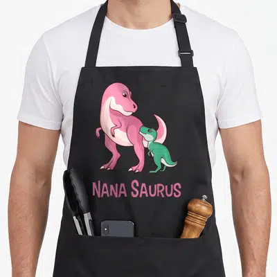 Saurus nana aprons - Pink and green cartoon dinosaurs standing above NANA SAURUS text — shown on a black cotton canvas apron against a white background