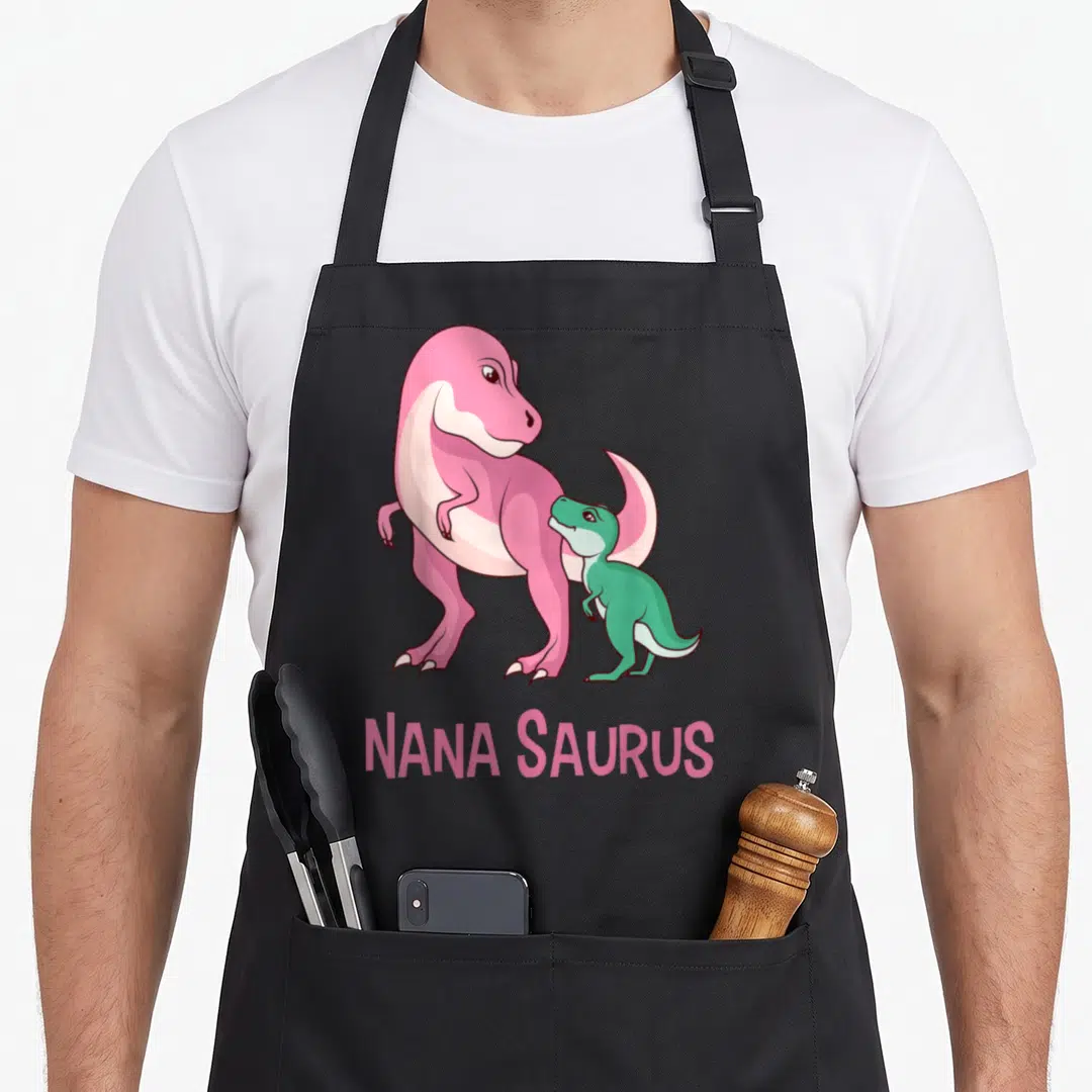 Saurus nana aprons - Pink and green cartoon dinosaurs standing above NANA SAURUS text — shown on a black cotton canvas apron against a white background