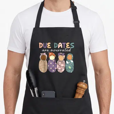 Due nurse apron - Four swaddled babies in peach, purple, teal, and white below due dates are overrated text — shown on a black cotton canvas apron against a white background
