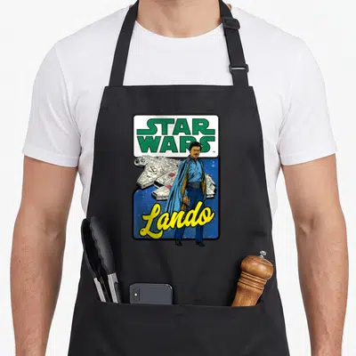 Lando falcon apron - Cartoon Lando Calrissian standing before the Millennium Falcon with yellow script text on a starry — shown on a black cotton canvas apron against a white background