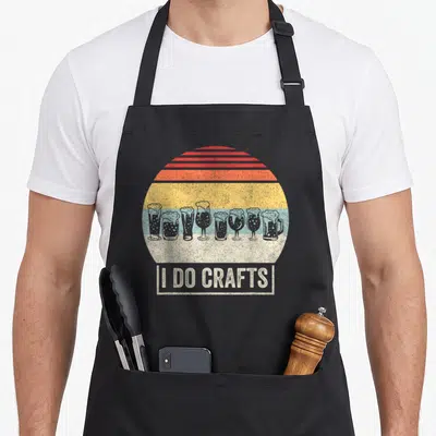 Brew beer craft apron - Seven beer glasses in retro striped sun circle above I DO CRAFTS text box — shown on a black cotton canvas apron against a white background