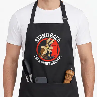 Stand professional aprons - Wile E. Coyote lighting red rocket with Stand Back I'm A Professional text in red and white — shown on a black cotton canvas apron against a white background