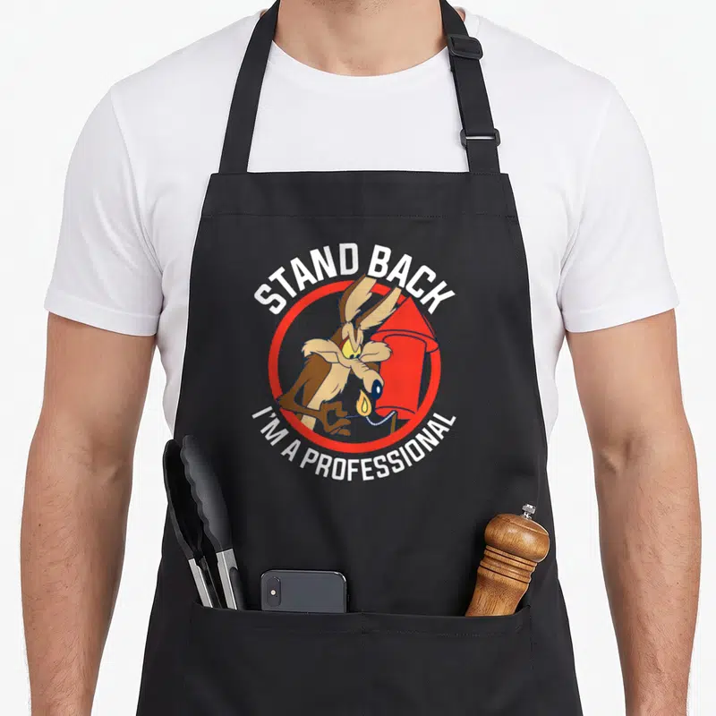 Stand professional aprons - Wile E. Coyote lighting red rocket with Stand Back I'm A Professional text in red and white — shown on a black cotton canvas apron against a white background