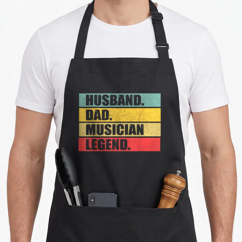 Musician father day apron - Stacked grey text reading husband dad musician legend on teal yellow beige and red — shown on a black cotton canvas apron against a white background