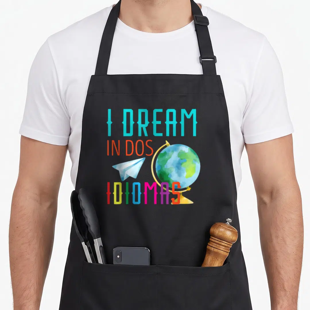 Dos teacher apron - I Dream in Dos Idiomas text with globe and paper plane in cyan red blue green yellow — shown on a black cotton canvas apron against a white background