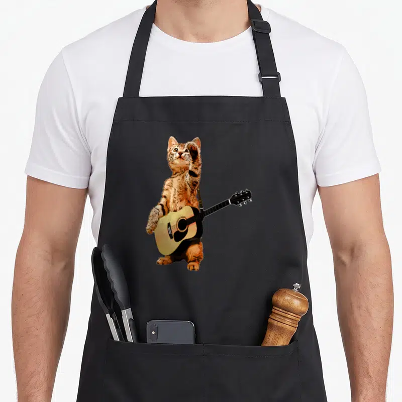 Musician cat apron - Orange tabby kitten standing playing acoustic guitar on black apron — shown on a black cotton canvas apron against a white background