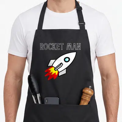 Spaceship arugula apron - ROCKET MAN text above a white cartoon rocket with red and yellow flames launching diagonally — shown on a black cotton canvas apron against a white background