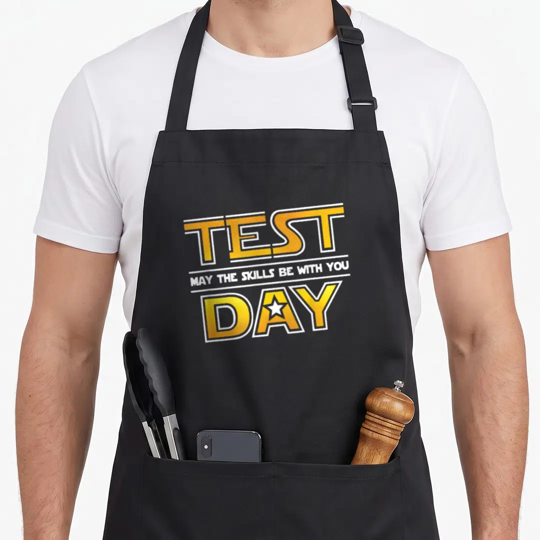May test kitchen apron - Yellow and white stacked text reading TEST DAY with a white star inside the letter — shown on a black cotton canvas apron against a white background