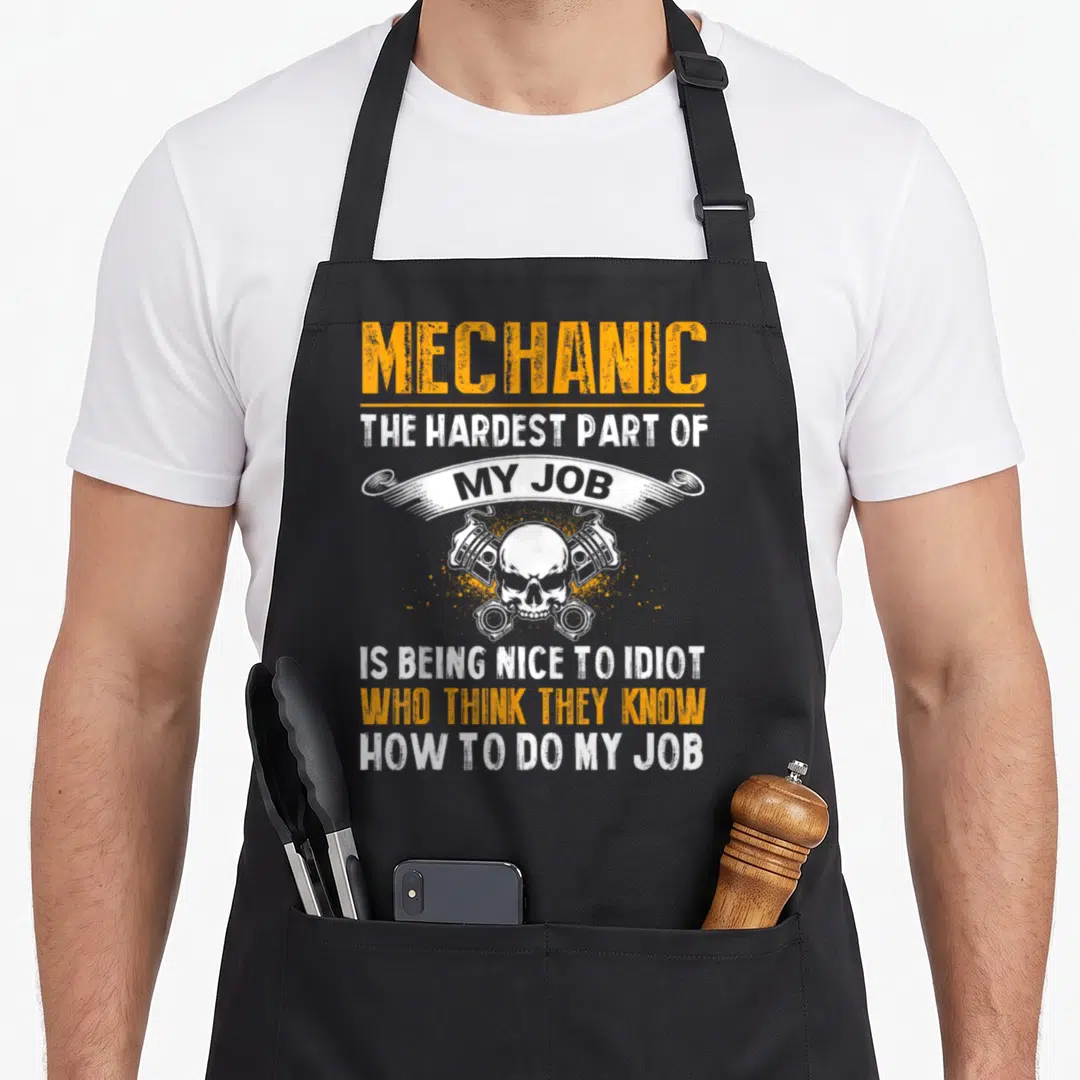 Hardest auto mechanic apron - Skull crossed with two pistons and sarcastic orange and white distressed text on gray — shown on a black cotton canvas apron against a white background