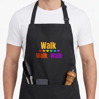 Walk outdoor apron - Walk text with six rainbow hearts and two lower Walk words on black fabric — shown on a black cotton canvas apron against a white background