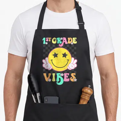 1st face apron - Yellow smiley face with star eyes, peace signs, 1ST GRADE VIBES text, checkerboard, flowers — shown on a black cotton canvas apron against a white background