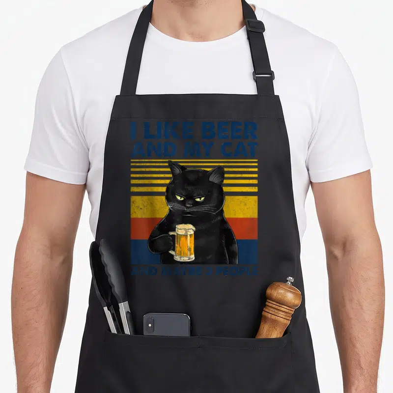 Beer cat apron - Black cat holding beer mug against yellow orange and blue stripes with text — shown on a black cotton canvas apron against a white background