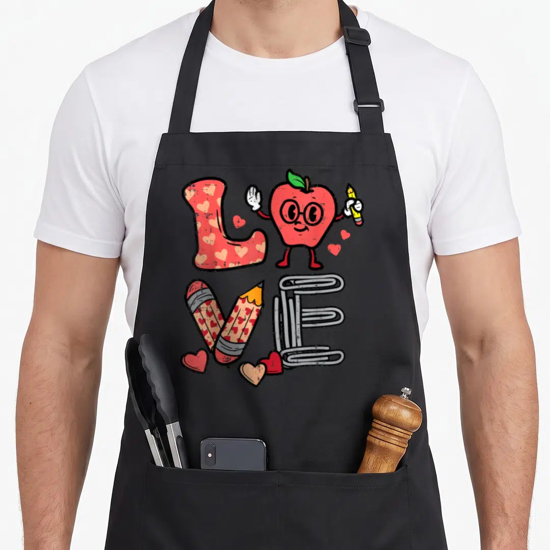 Apple valentine's day apron - Cartoon apple and school supplies arranged to spell love in red grey white yellow — shown on a black cotton canvas apron against a white background