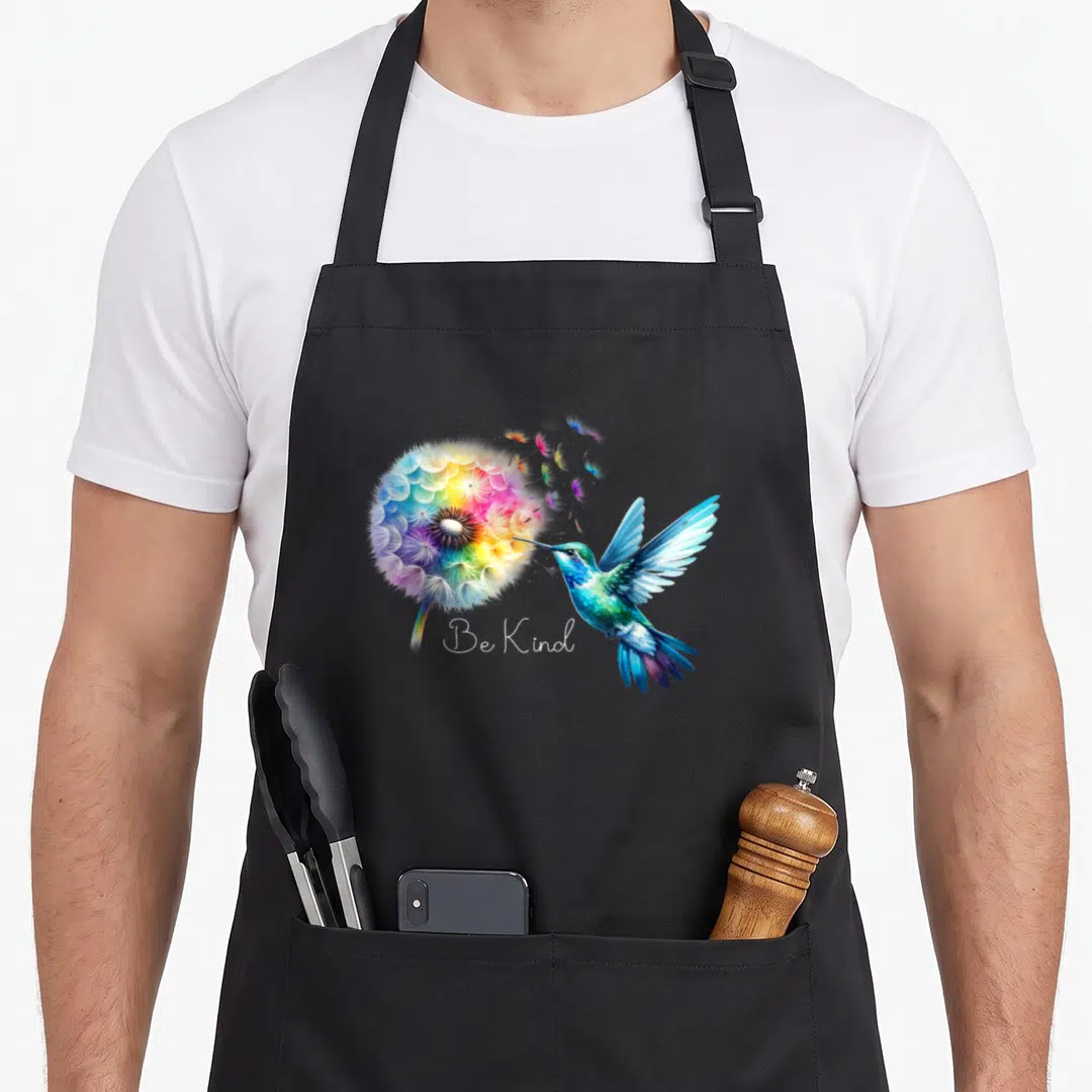 Dandelion the floral apron - Rainbow dandelion and blue hummingbird watercolor art with Be Kind text — shown on a black cotton canvas apron against a white background