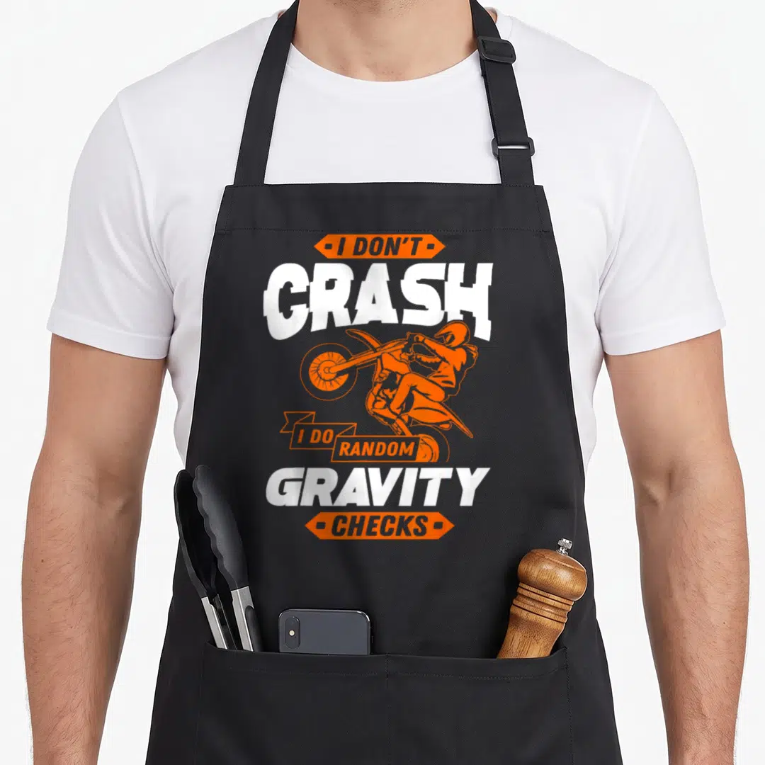 Random bike apron - I Don't Crash I Do Random Gravity Checks text with orange jumping motocross rider silhouette — shown on a black cotton canvas apron against a white background