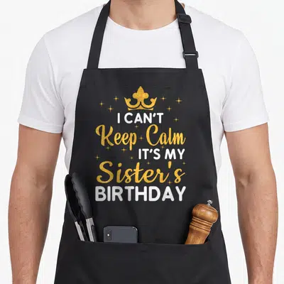 Funny sister birthday apron - Gold crown and white text I Can't Keep Calm It's My Sister's Birthday with stars — shown on a black cotton canvas apron against a white background