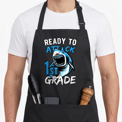 Attack school apron - Cartoon shark with open mouth surrounded by ready to attack 1st grade text in blue and white — shown on a black cotton canvas apron against a white background