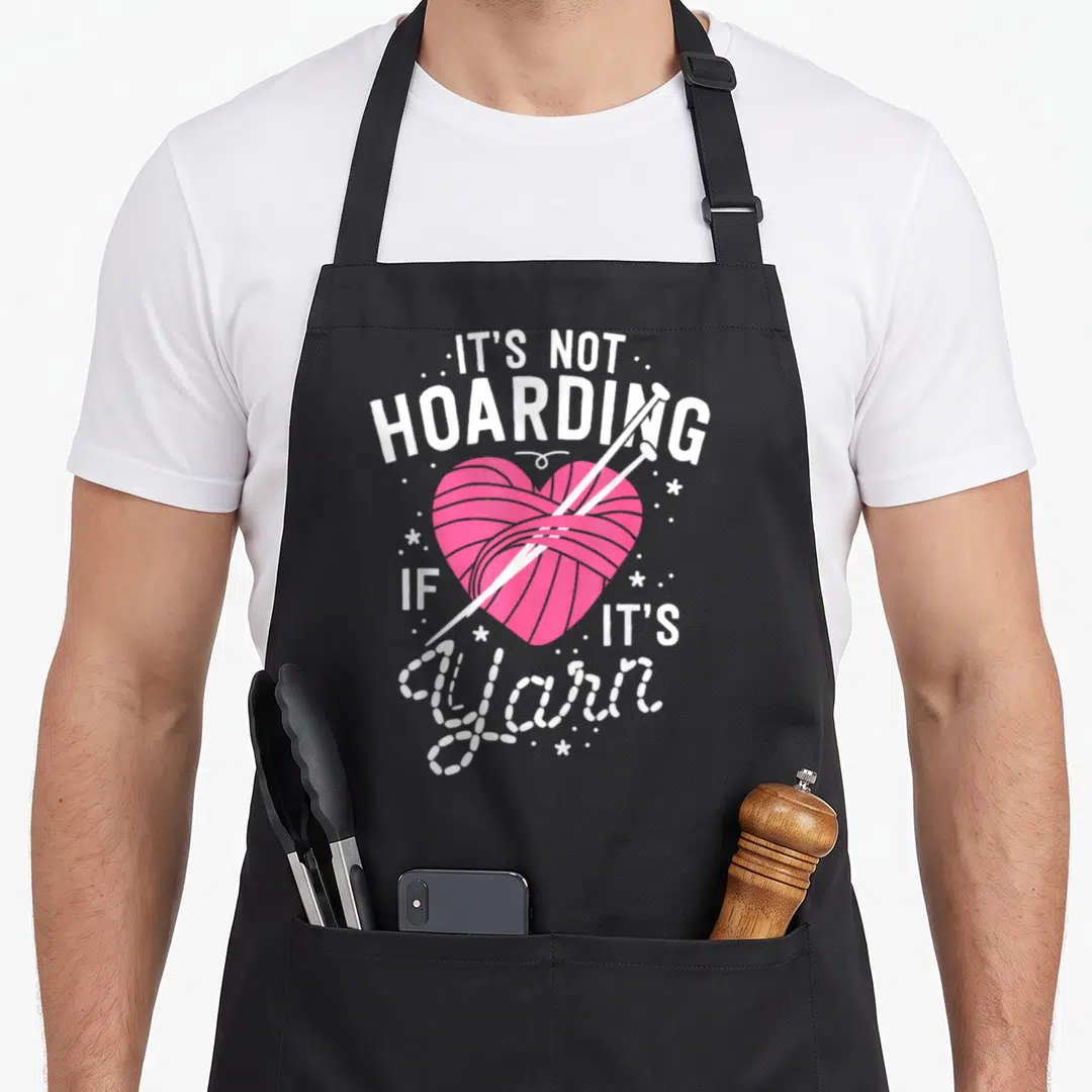 Hoarding yarn knitting apron - Pink heart yarn ball with crossed needles and white dotted script text — shown on a black cotton canvas apron against a white background