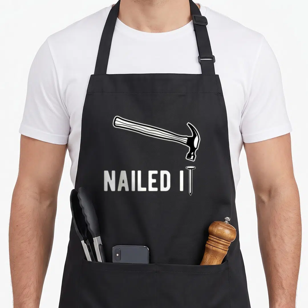 Nailed woodworking apron - Black and white hammer graphic above NAILED IT text with nail replacing the letter I — shown on a black cotton canvas apron against a white background