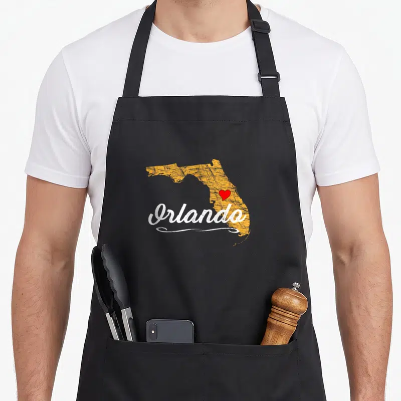 Orlando vacation souvenir florida apron - Distressed orange Florida map with white Orlando script and red heart marker — shown on a black cotton canvas apron against a white background