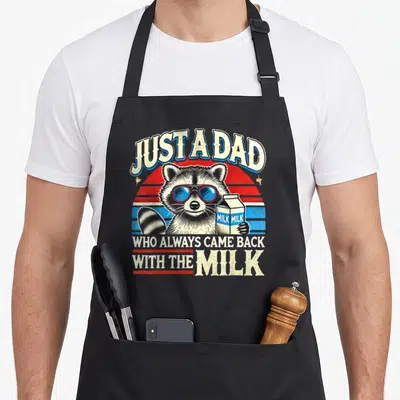 Racoon milk aprons - Retro raccoon in sunglasses holding a milk carton under arched text JUST A DAD — shown on a black cotton canvas apron against a white background