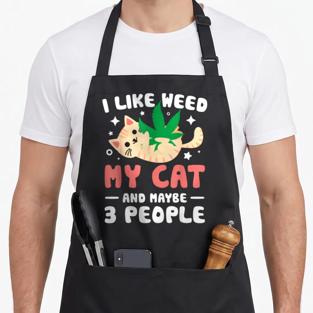 Weed cat apron - Cartoon ginger cat holding green leaf with stacked text I LIKE WEED MY CAT AND MAYBE 3 PEOPLE — shown on a black cotton canvas apron against a white background