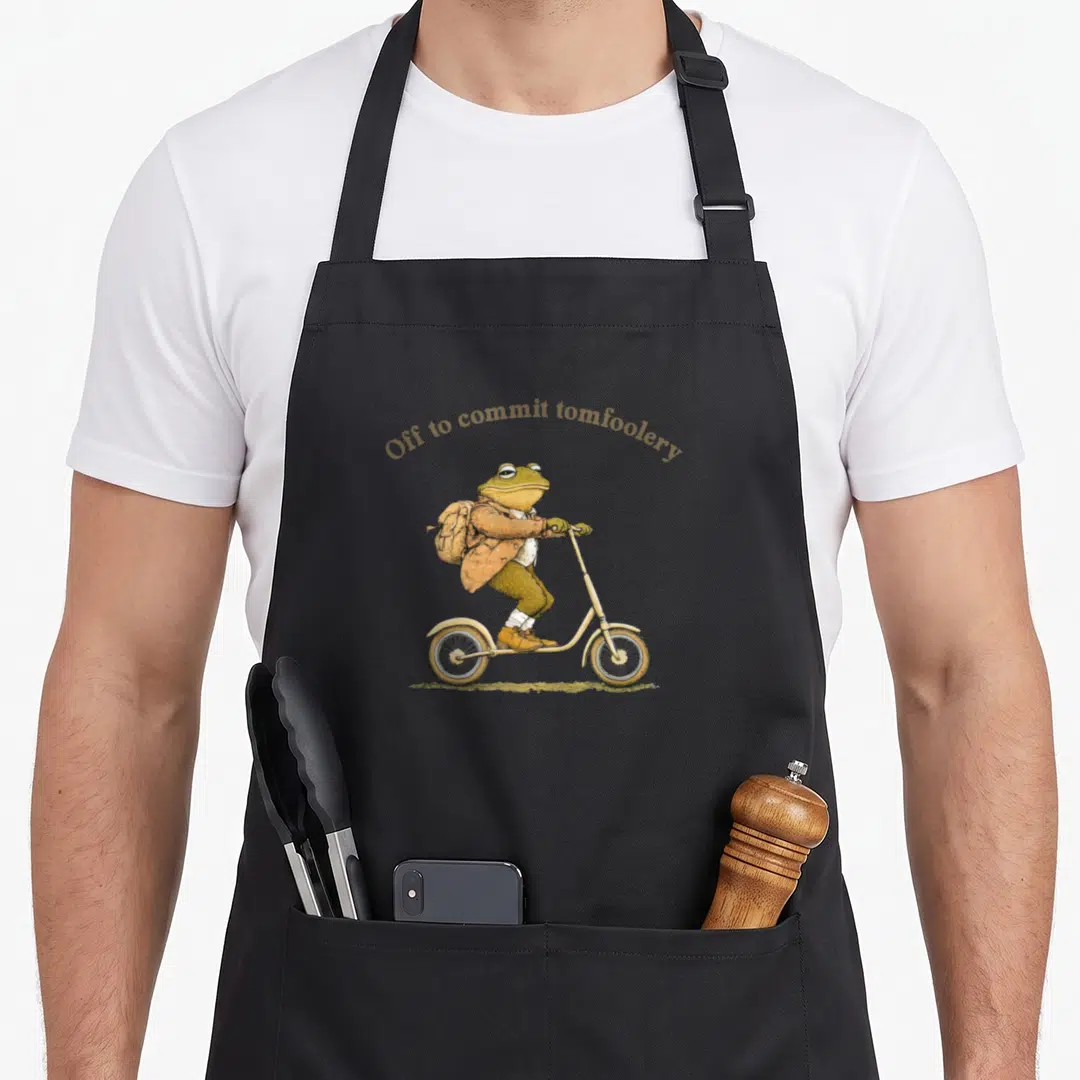 Toad cottagecore apron - Frog in clothes riding scooter under arched text Off to commit tomfoolery in gold and green — shown on a black cotton canvas apron against a white background