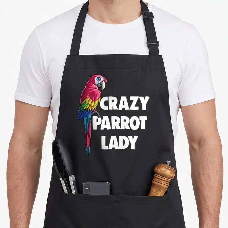 Cool crazy parrot lady apron - Cartoon parrot with pink and blue feathers beside stacked white text reading CRAZY — shown on a black cotton canvas apron against a white background