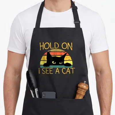 See cat apron - Retro sunset with black cat head paws and text Hold On I See A Cat — shown on a black cotton canvas apron against a white background