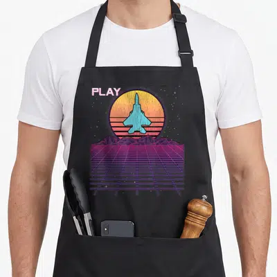 Raptor f18 apron - Retro fighter jet over striped sun with PLAY text and purple grid landscape — shown on a black cotton canvas apron against a white background