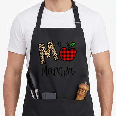Maestra apple apron - Leopard M and red plaid apple with M de Maestra text — shown on a black cotton canvas apron against a white background