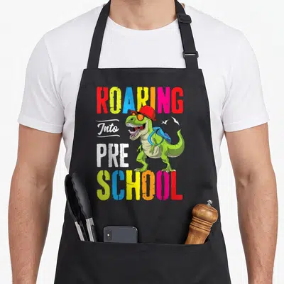 Roaring preschool teacher apron - Green dinosaur with red cap, blue backpack, and ROARING Into PRE SCHOOL text — shown on a black cotton canvas apron against a white background
