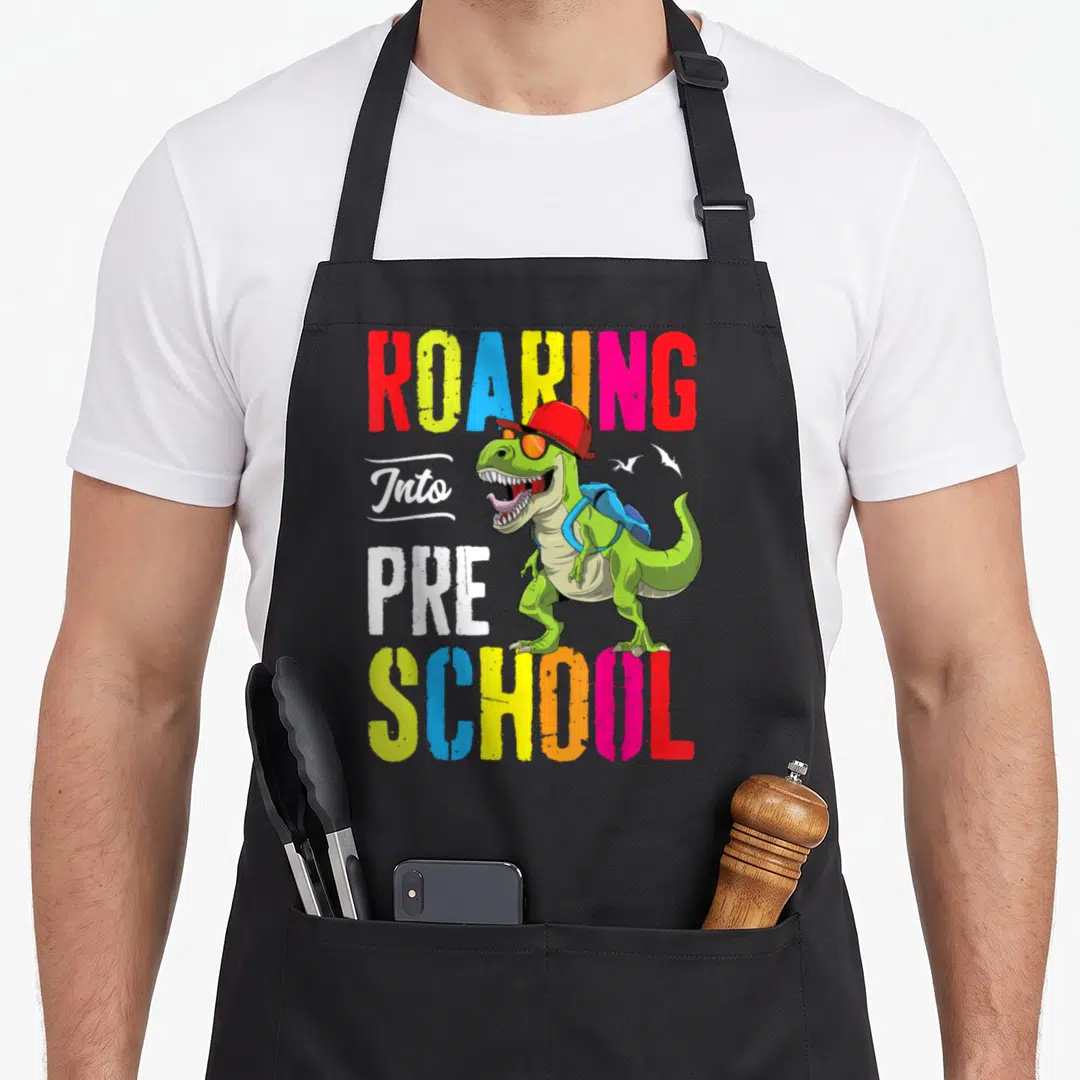 Roaring preschool teacher apron - Green dinosaur with red cap, blue backpack, and ROARING Into PRE SCHOOL text — shown on a black cotton canvas apron against a white background