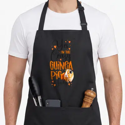 Animal obsessed guinea lady apron - Stacked text reading Yes Im That Crazy Guinea Pig Lady with a tricolor guinea pig — shown on a black cotton canvas apron against a white background