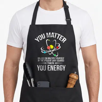 Matter geek aprons - Cartoon atom model with multicolored electrons and distressed white text about matter and energy — shown on a black cotton canvas apron against a white background