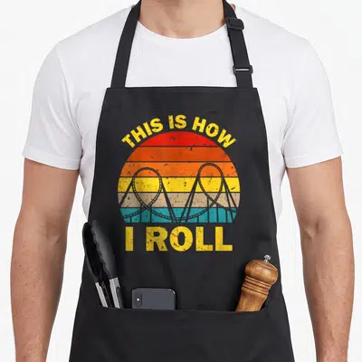 Roller theme aprons - Retro striped sun with grey roller coaster loops and yellow THIS IS HOW I ROLL text — shown on a black cotton canvas apron against a white background