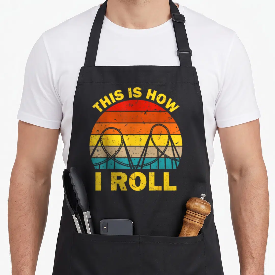 Roller theme aprons - Retro striped sun with grey roller coaster loops and yellow THIS IS HOW I ROLL text — shown on a black cotton canvas apron against a white background