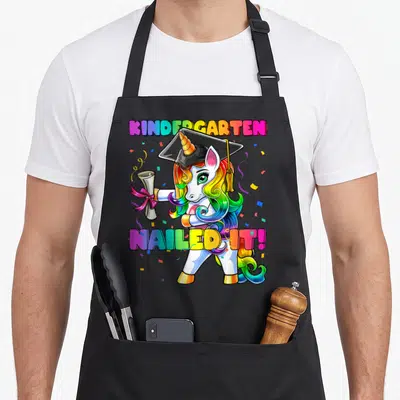 Flossing unicorn aprons - Cartoon unicorn in cap holding diploma with text KINDERGARTEN NAILED IT! — shown on a black cotton canvas apron against a white background