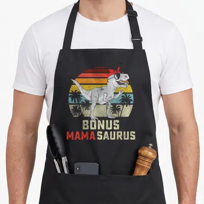 Bonus family aprons - T-Rex in sunglasses and red bandana with BONUS MAMA SAURUS text over striped retro sun — shown on a black cotton canvas apron against a white background