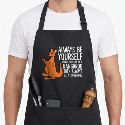 Alway kangaroo apron - Orange cartoon kangaroo standing next to stacked white text reading Always Be Yourself Unless — shown on a black cotton canvas apron against a white background