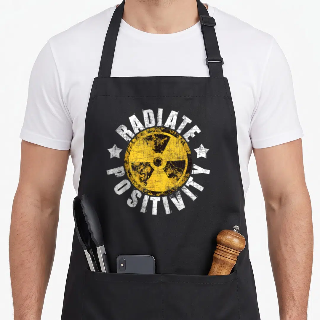 Radiate radiology apron - Distressed radiation symbol with RADIATE POSITIVITY text and white stars — shown on a black cotton canvas apron against a white background
