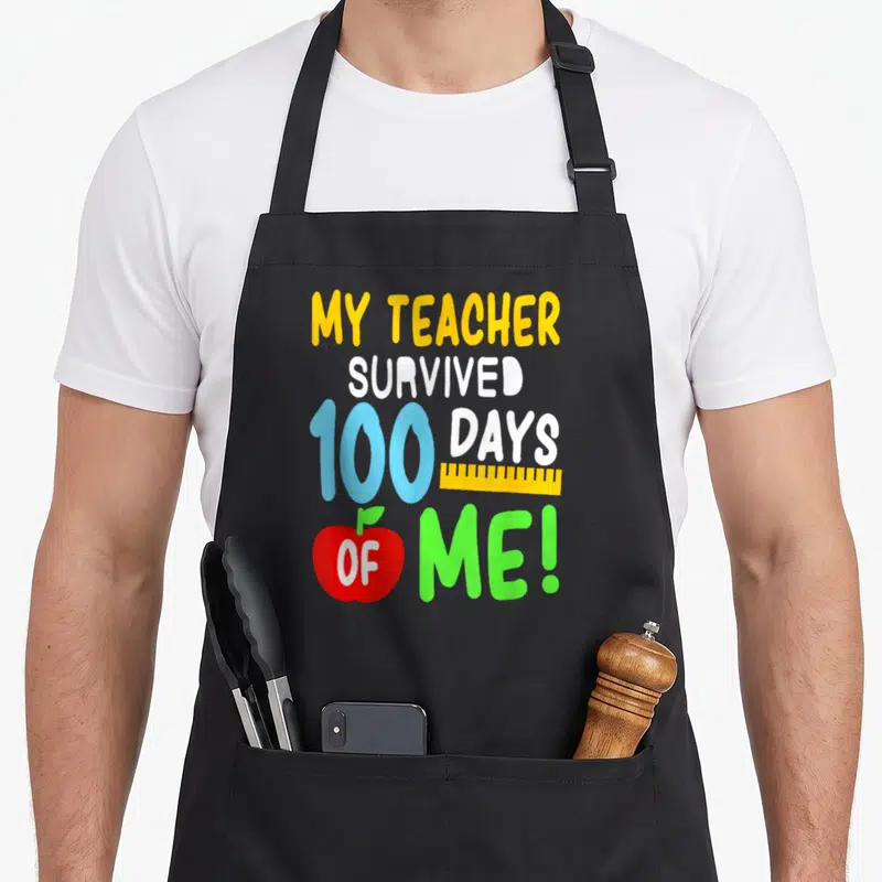 Survived school teacher apron - Stacked colorful text My Teacher Survived 100 Days Of Me with yellow ruler and red — shown on a black cotton canvas apron against a white background