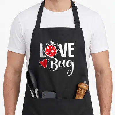 Valentine ladybug apron - Red ladybug replacing O in white LOVE text with red heart next to Bug script — shown on a black cotton canvas apron against a white background