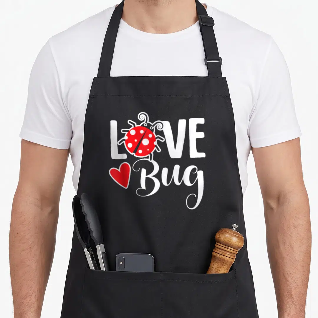 Valentine ladybug apron - Red ladybug replacing O in white LOVE text with red heart next to Bug script — shown on a black cotton canvas apron against a white background