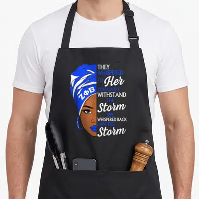 Zeta african apron - Woman portrait with ZFB headwrap and storm quote text in blue and white — shown on a black cotton canvas apron against a white background