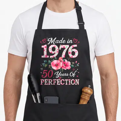50th birthday vintage floral apron - Pink poppies and floral text reading Made in 1976 50 Years Of Perfection — shown on a black cotton canvas apron against a white background