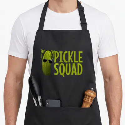 Funny cucumber pickle apron - Cartoon pickle wearing sunglasses beside lime green PICKLE SQUAD text — shown on a black cotton canvas apron against a white background